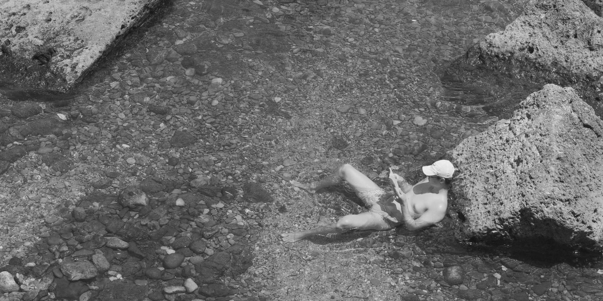 Person reading in shallow water beside rocks, a moment of quiet focus and relaxation