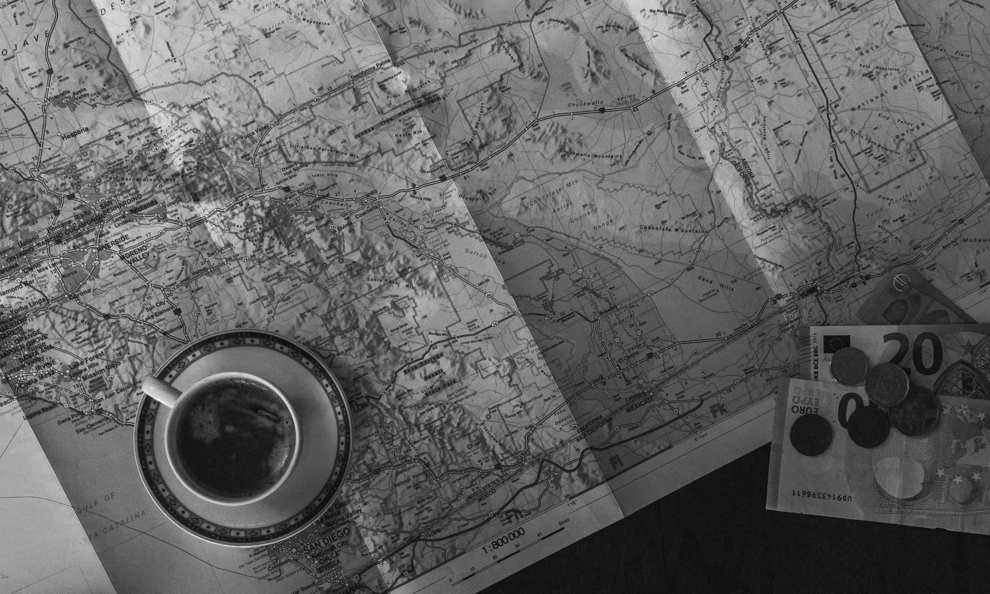 Black-and-white travel flat lay with a folded map, a cup of coffee, and euro notes