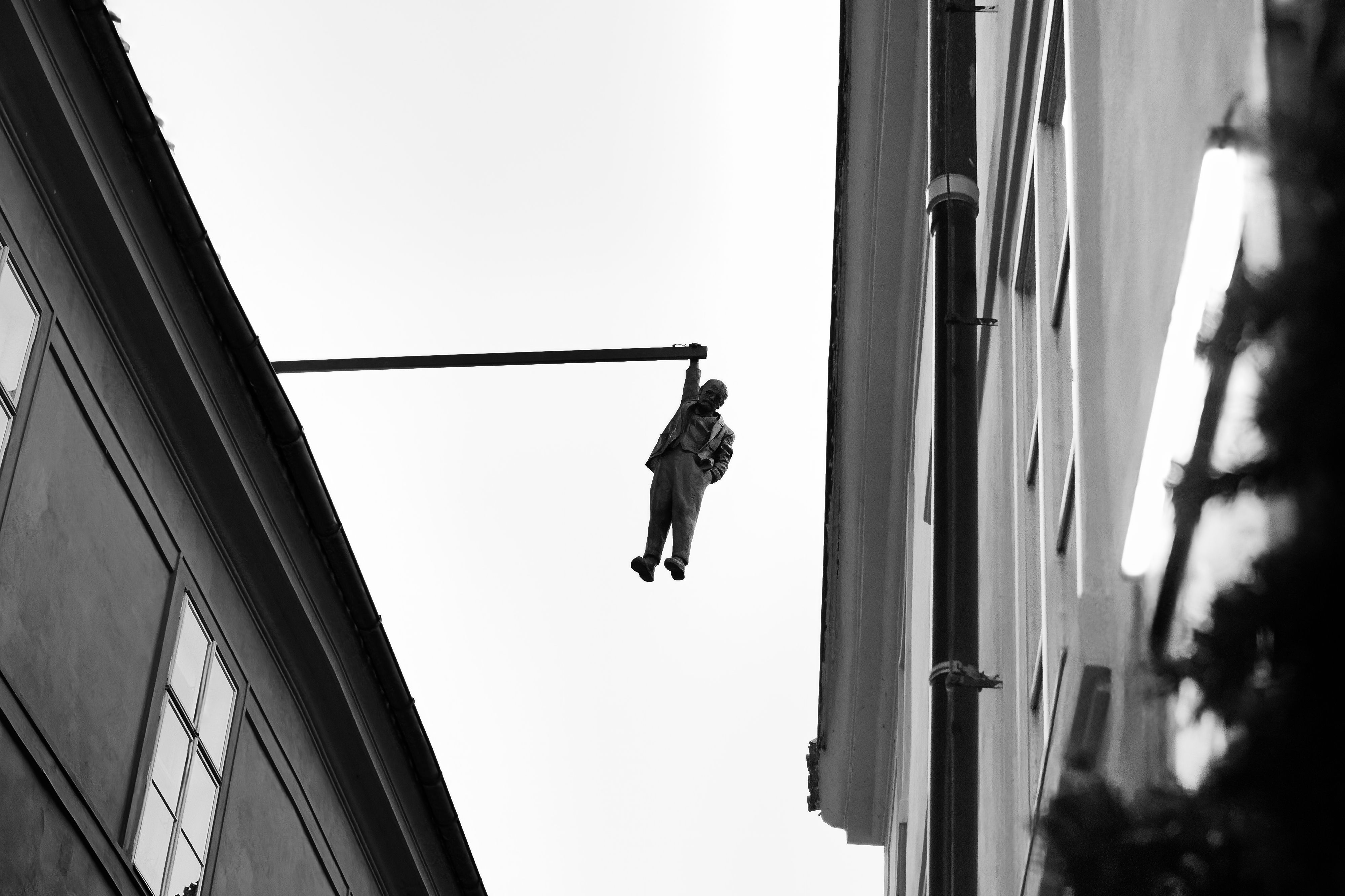Black-and-white view looking up at David Černý's Man Hanging Out sculpture suspended between two buildings